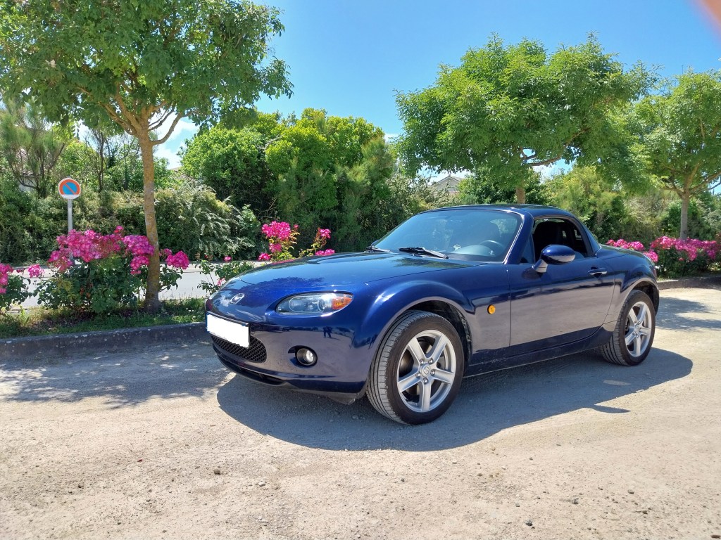 Mazda MX-5 NC bleue en mode coupé garée sur parking.