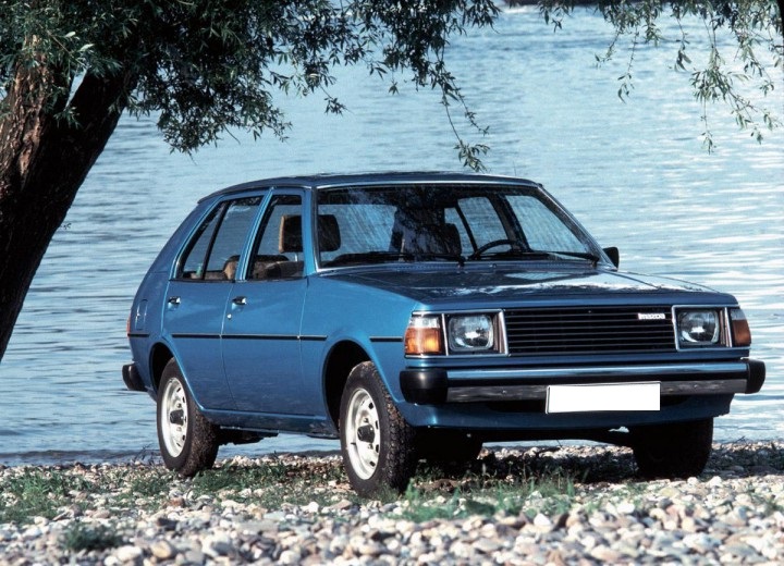 Mazda 323 FA bleue sous un arbre devant un lac.